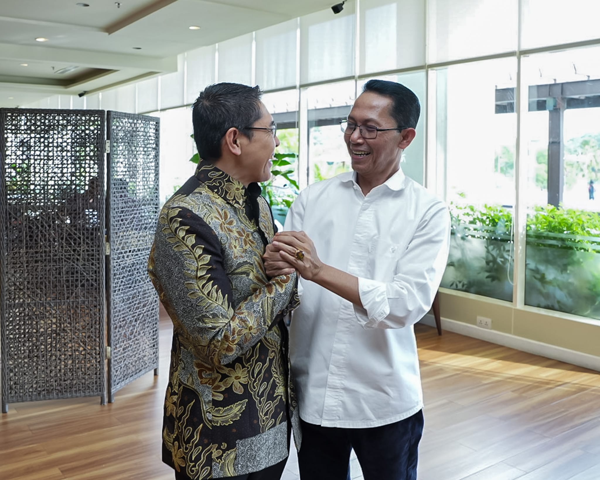 Two men with glasses shaking hands in a brightly lit room with a brown woven screen.